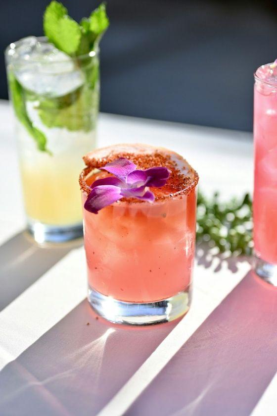 Bunte Cocktails auf einem Tisch, darunter ein pinker Drink mit einer Blüte als Garnitur, serviert bei einem Afterwork-Cocktailabend auf der Dachterrasse.
